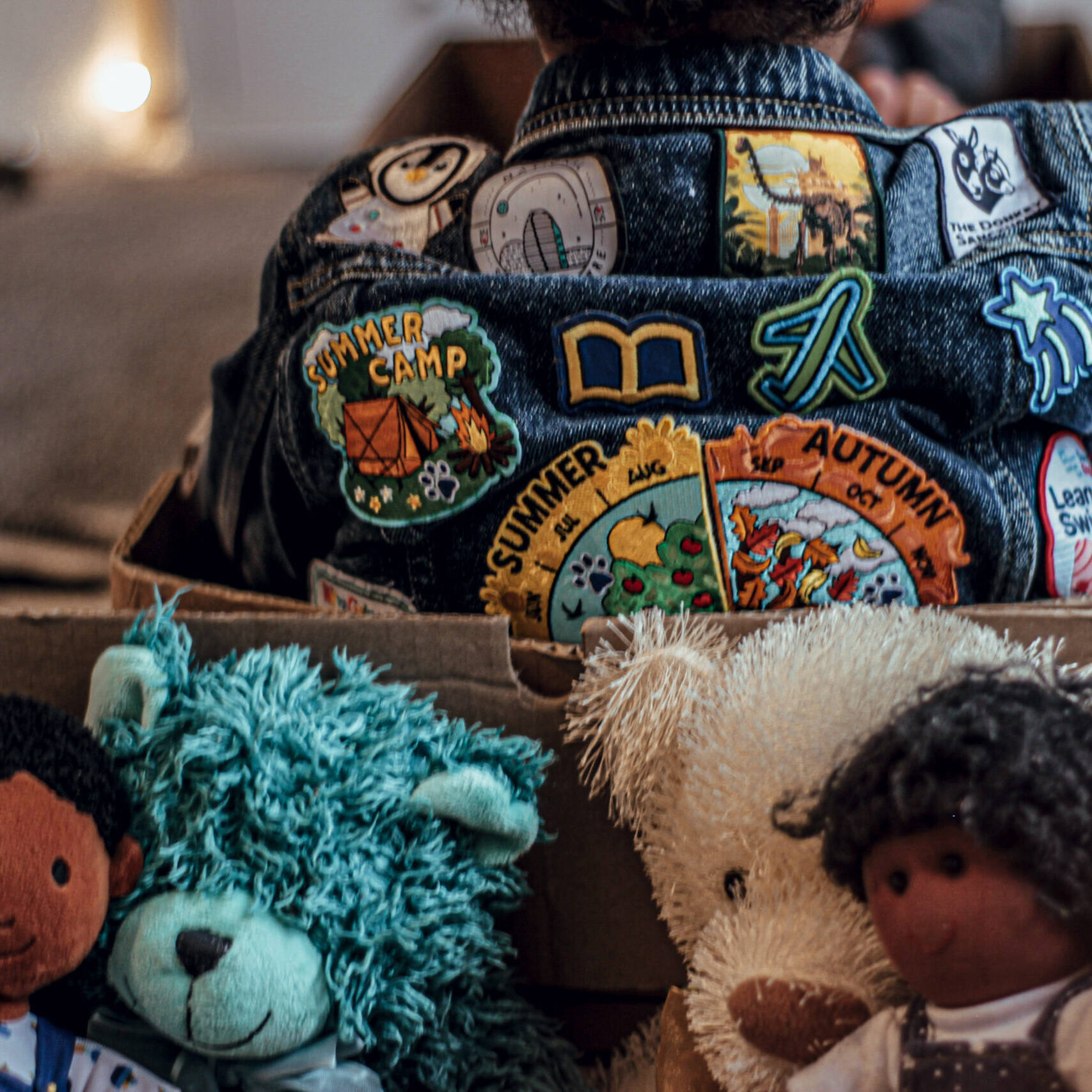 A Kid Sitting In A Box With The Back Part Of His Jacket Showing At The Top. It Has Several Badges Sewn On It. There are Four Teddies in A Box Too.