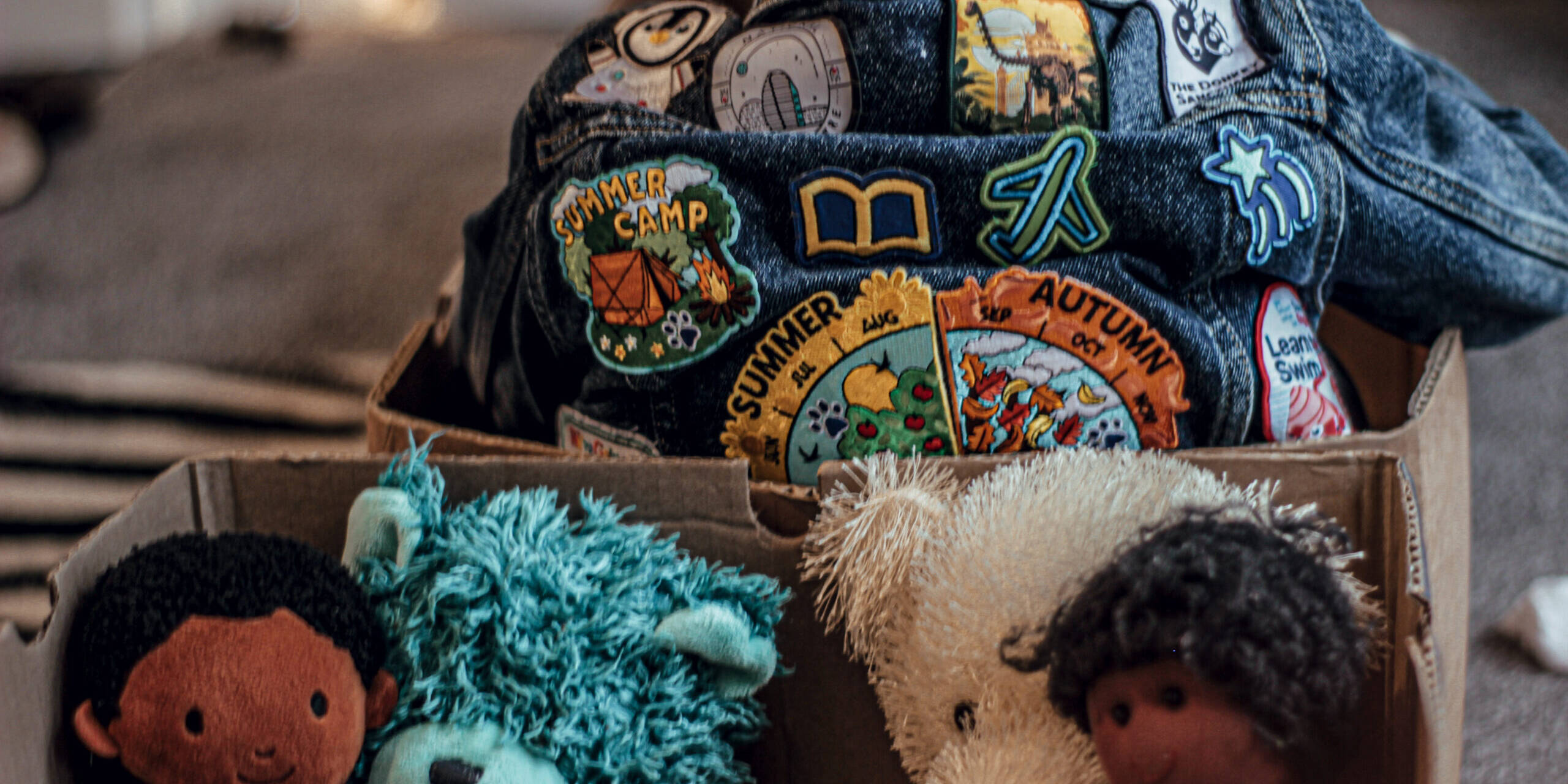 A Kid Sitting In A Box With The Back Part Of His Jacket Showing At The Top. It Has Several Badges Sewn On It. There are Four Teddies in A Box Too.