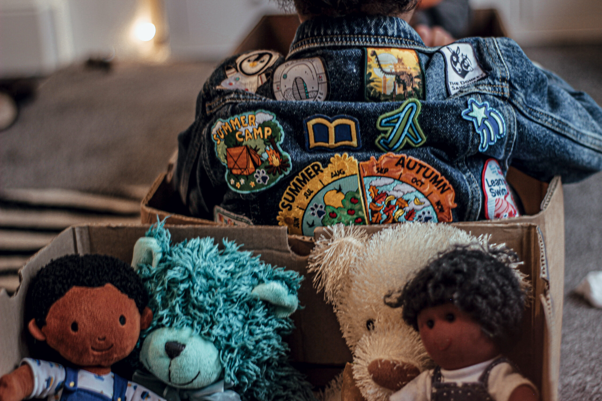 A Kid Sitting In A Box With The Back Part Of His Jacket Showing At The Top. It Has Several Badges Sewn On It. There are Four Teddies in A Box Too.